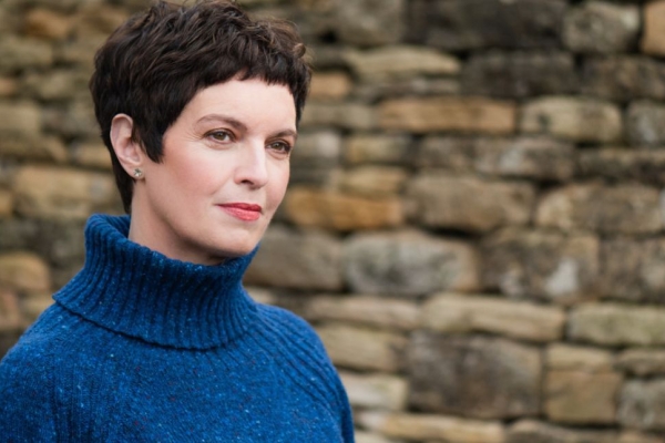 A colour portrait of Louise Tickle in front of a stone wall. Tickle is a white woman with short dark hair wearing a blue polo necked jumper. She faces at angle to the camera.